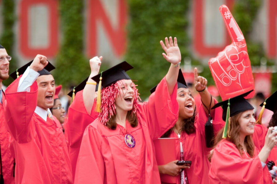 Photo: A group of graduating students all wearing red graduation gowns and black caps, cheer and shout. One student wears a red pom pom under their cap.