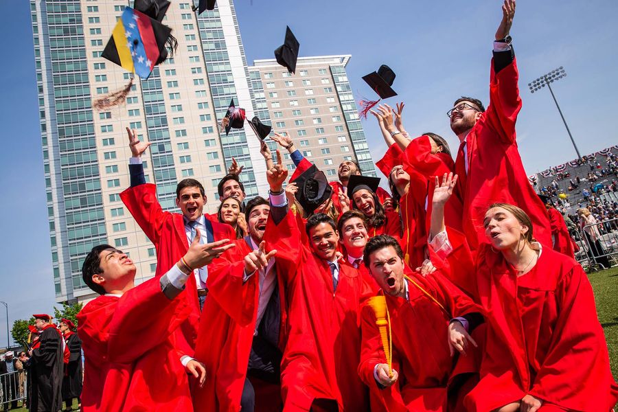 Photo: A group of students wearing red graduation gowns and black caps celebrate and cheer on Nickerson Field. A few of them throw their caps into the air as they cheer.