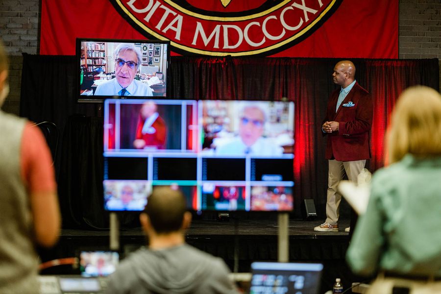 Photo: 5 large screens are shown on stage as Dean Elmore, a Black man wearing a light blue collared shirt, red velvet jacket, and tan pants looks on to one screen that shows the large face of Robert Pinsky, an older white man shown in his home office.