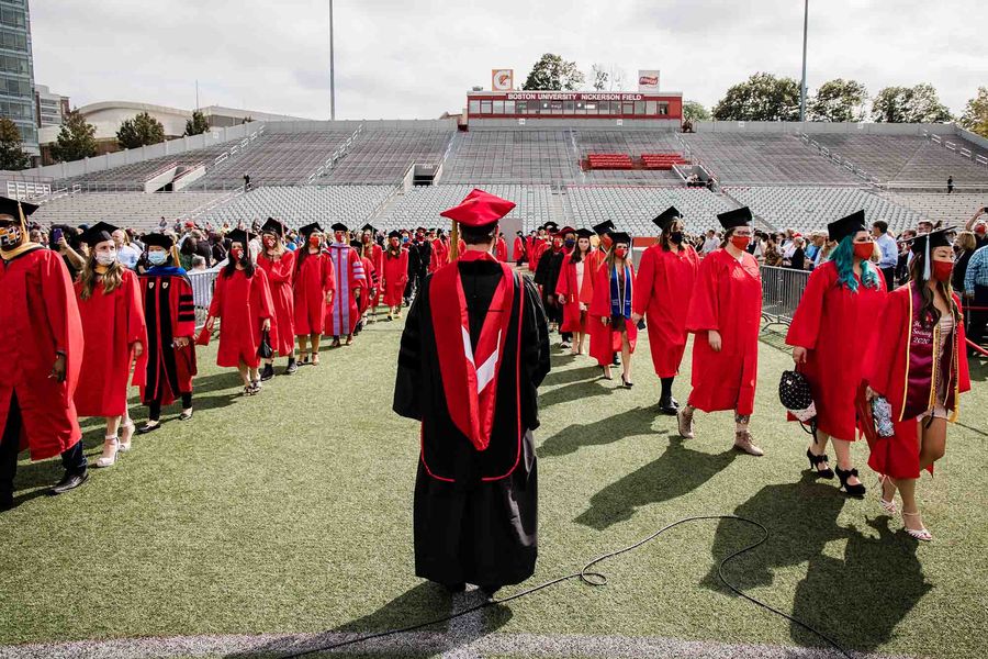 Photo: Students wearing red graduation gowns, black caps, and red face masks file into Nickerson Field for commencement. A center person wearing a black gown and red cap stands in the center facing away from the camera.