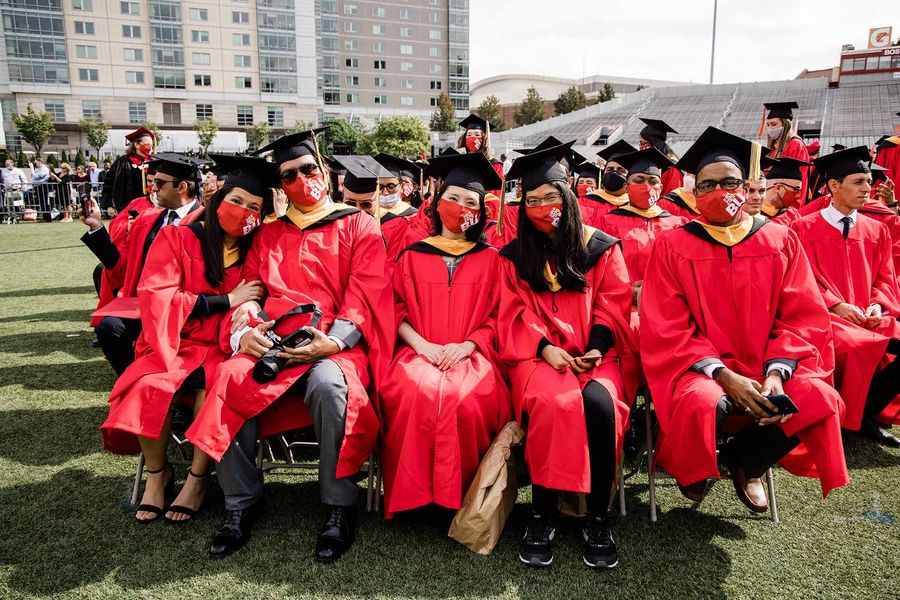 Photo. A group of students sit on chairs on Nickerson Field. They all wear red gowns, black graduation caps, a red face masks that read "BU" in large white letters.