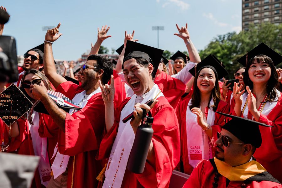 Photo: A large group shot of graduating students wearing red gowns and black graduation caps celebrating and cheering loudly.