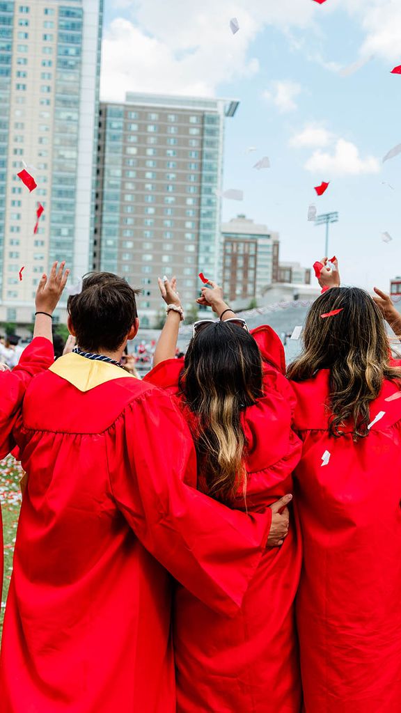 Photo: Shot of a group of graduating students wearing red gowns facing away from the camera and celebrating. Red and white confetti falls from the sky and Stuvi II can be seen in the distance.