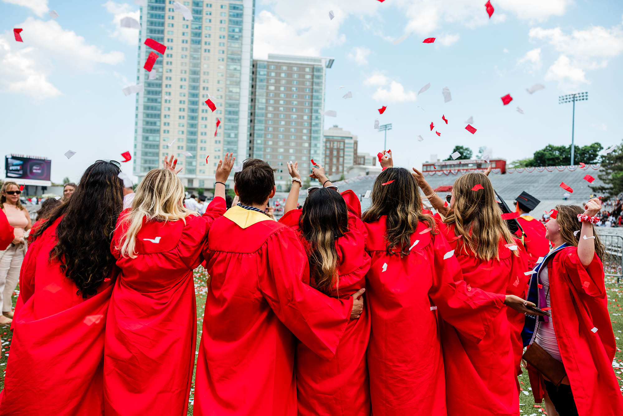 Photo: Shot of a group of graduating students wearing red gowns facing away from the camera and celebrating. Red and white confetti falls from the sky and Stuvi II can be seen in the distance.
