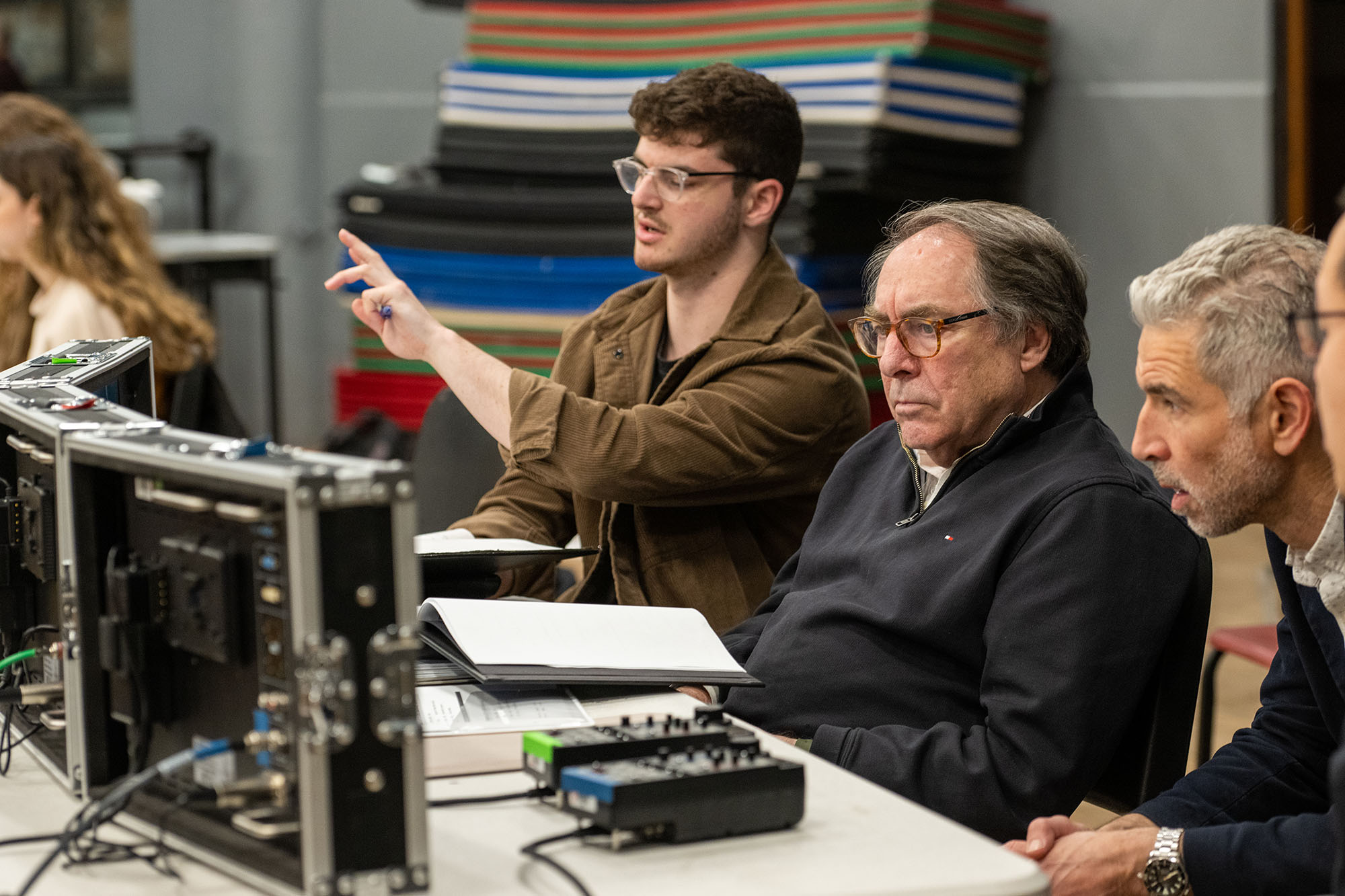 Photo: From left, director Eli Canter (COM’23), Paul Schneider, COM professor of film and television and department chair, and Tim Palmer, a COM professor of the practice in cinematography, discuss camera blocking during a rehearsal. A young man wearing glasses and a brown collared shirt points forward and speaks as an older man wearing glasses and a black collared shirt sits to his right. They both sit at a table with multiple mini screens in front of them.