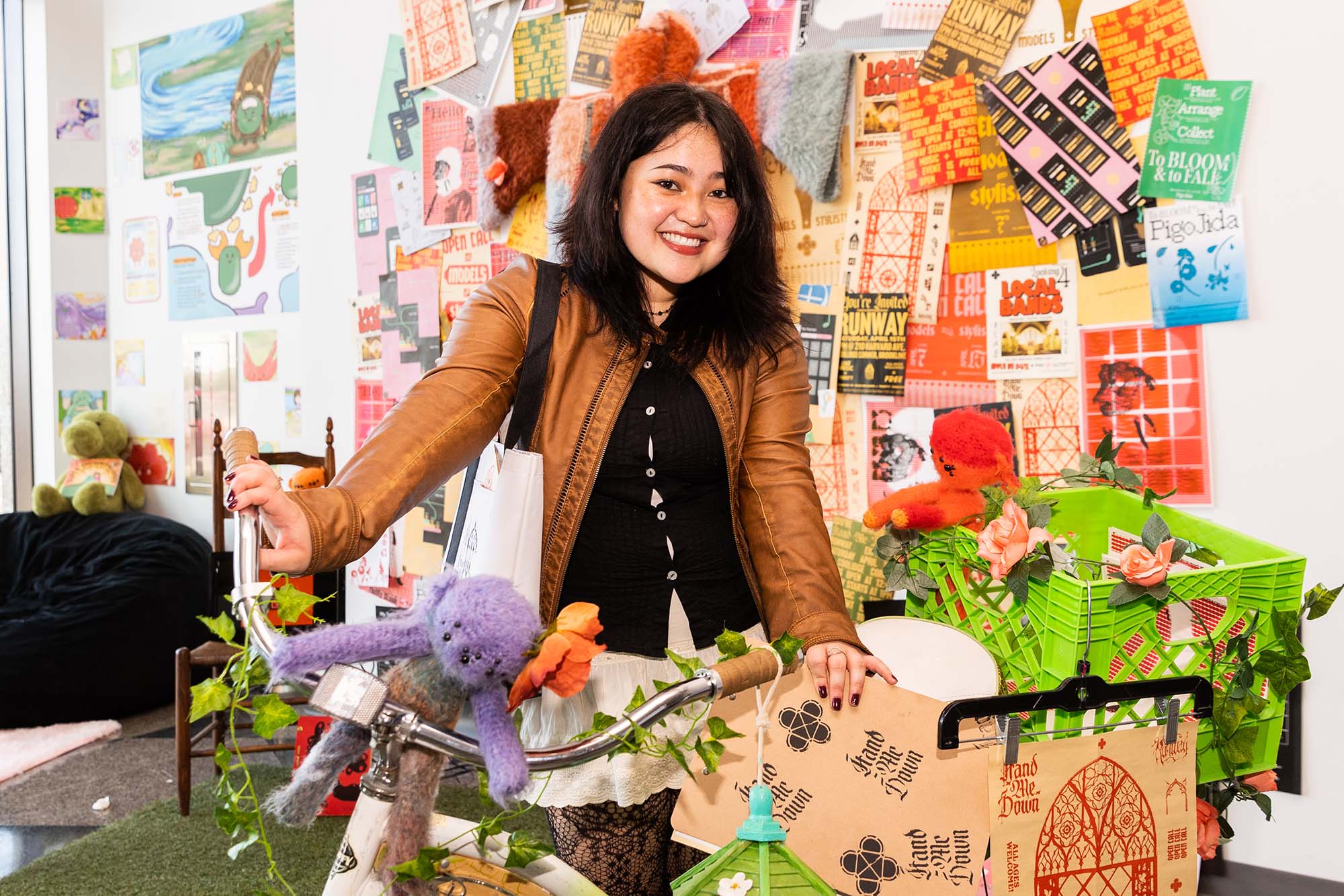 Photos: August Ramos (CFA’23) shows off some of the ephemera seen in her senior thesis runway show, “HAND-ME-DOWN". A young woman with shoulder-length black hair and wearing a black button-up collared shirt and tan jacket smiles and poses in front of a large #D collage made up of various items, articles, clippings, and bike handles.