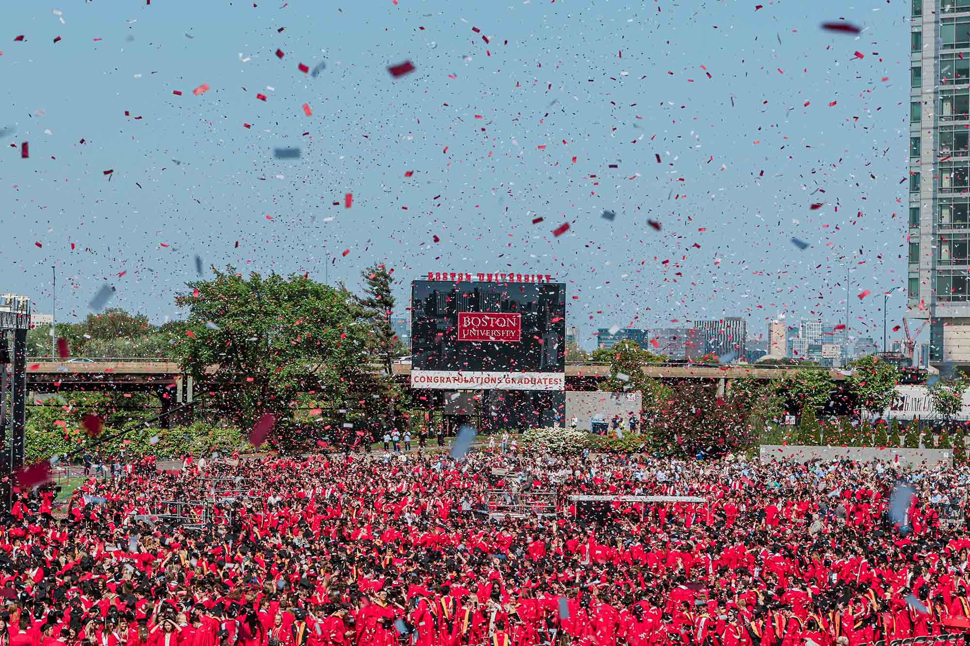 Photo: Graduates wearing red gowns and black graduation caps cheer and celebrate at the end of the 150th Boston University Commencement on Nickerson Field as red and white confetti falls from the air onto them.