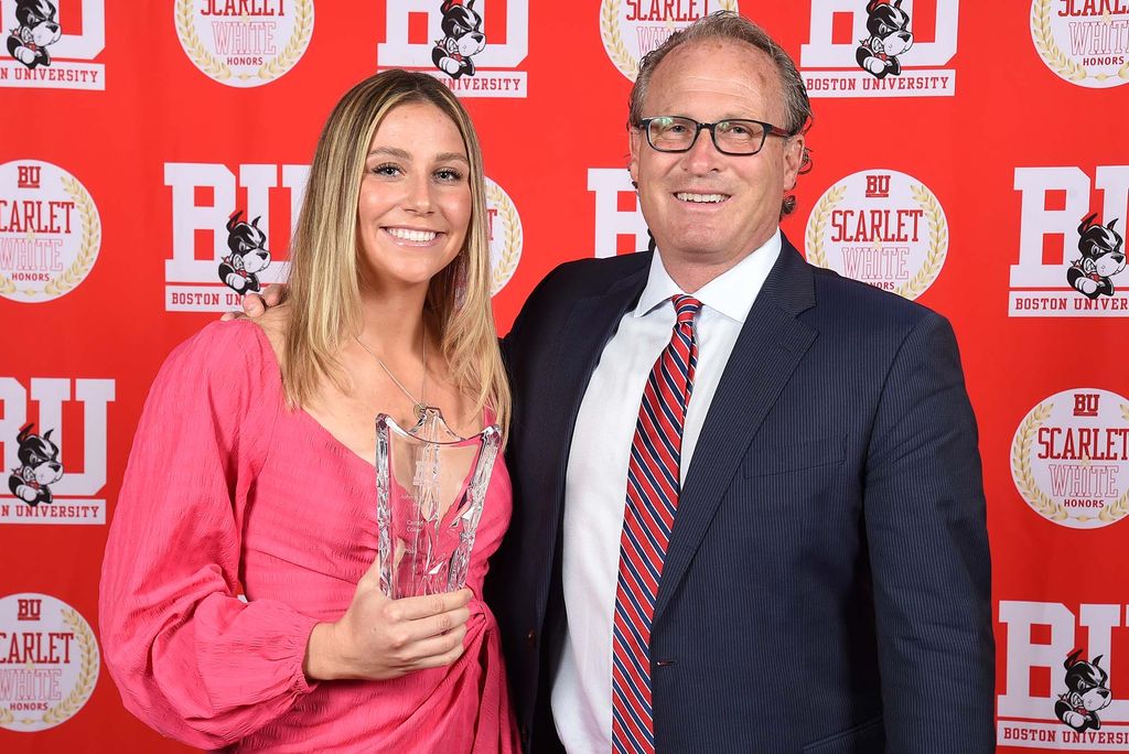 Photo: Caitlin Coker, a young white woman in a pink dress stands next to an older man in a suit, in front of a red BU photo background