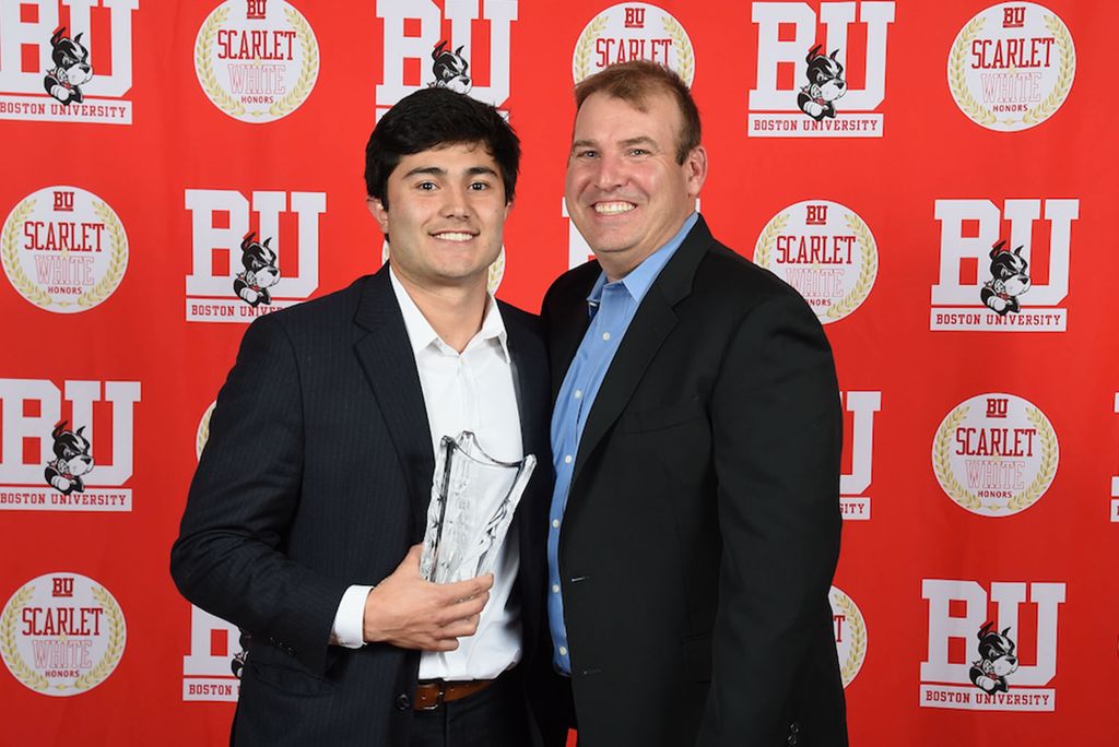 Photo: Roy Meyer a young white man in a suit stands next to an older man in a suit, in front of a red BU photo background