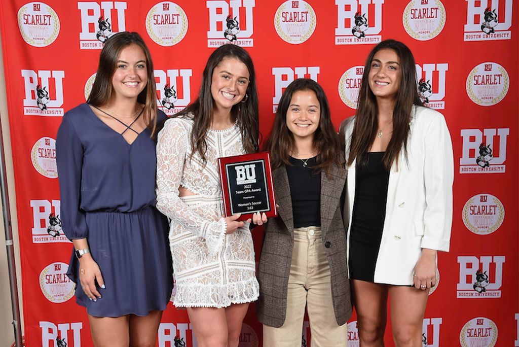 Photo: 4 young women, members of the BU women's soccer team, hold an award that they received while standing in front of a red BU background