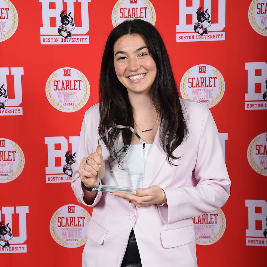 Photo: A young woman with dark hair in a light blazer holds up an award in front of a red background