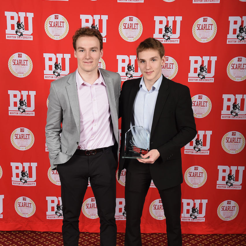 Photo: Two young men with dark hair in suits hold up an award in front of a red background