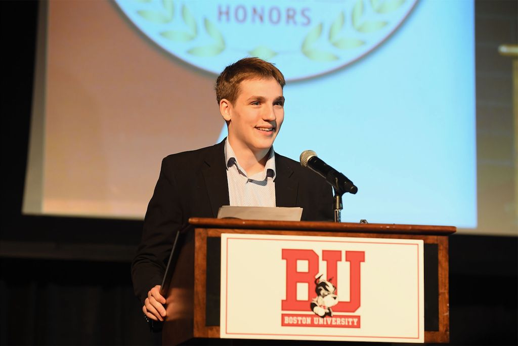 Photo: Lane Hutson, a young white man wearing a suit stands at a podium after accepting an award for Ice Hockey