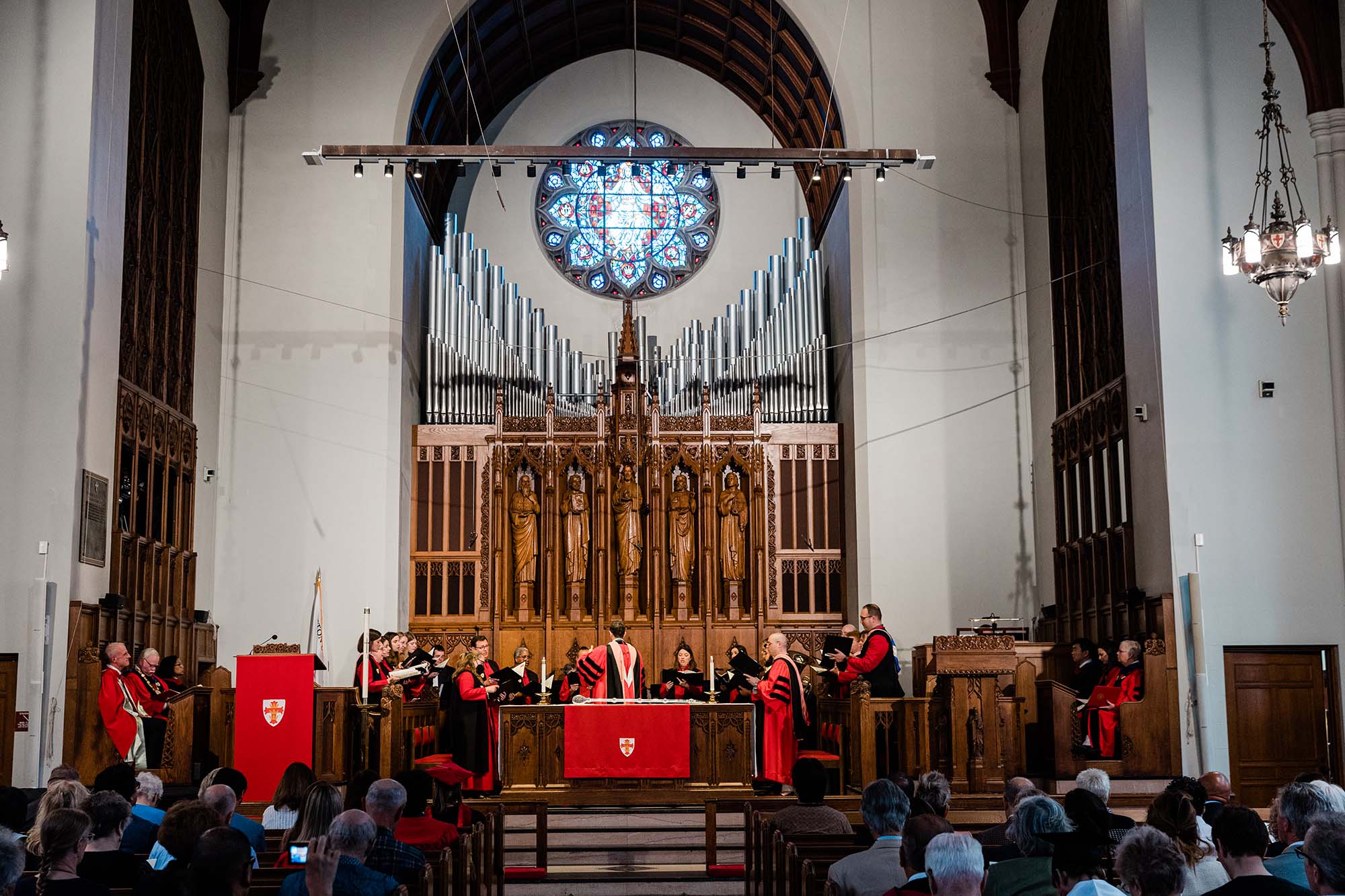 Photo: wide shot of the 150th Boston University Baccalaureate service at Marsh Chapel. People sit in the audience and watch as the choir, dressed in red and black ceremonial robes, sing. Ornate windows and a large organ are shown behind them.