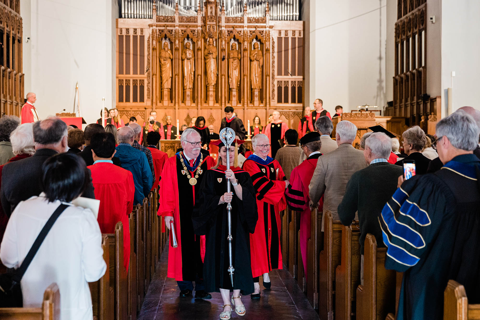 Photo: University President Robert A. Brown, an older white man wearing a red ceremonial gown (left),and provost and chief academic officer Jean Morrison (right), an older white woman wearing a red ceremonial gown, lead the recessional from Marsh Chapel at the end of the Baccalaureate service. Kimberly Howard, a white woman wearing wearing a black ceremonial gown and cap(center), carries the University mace.