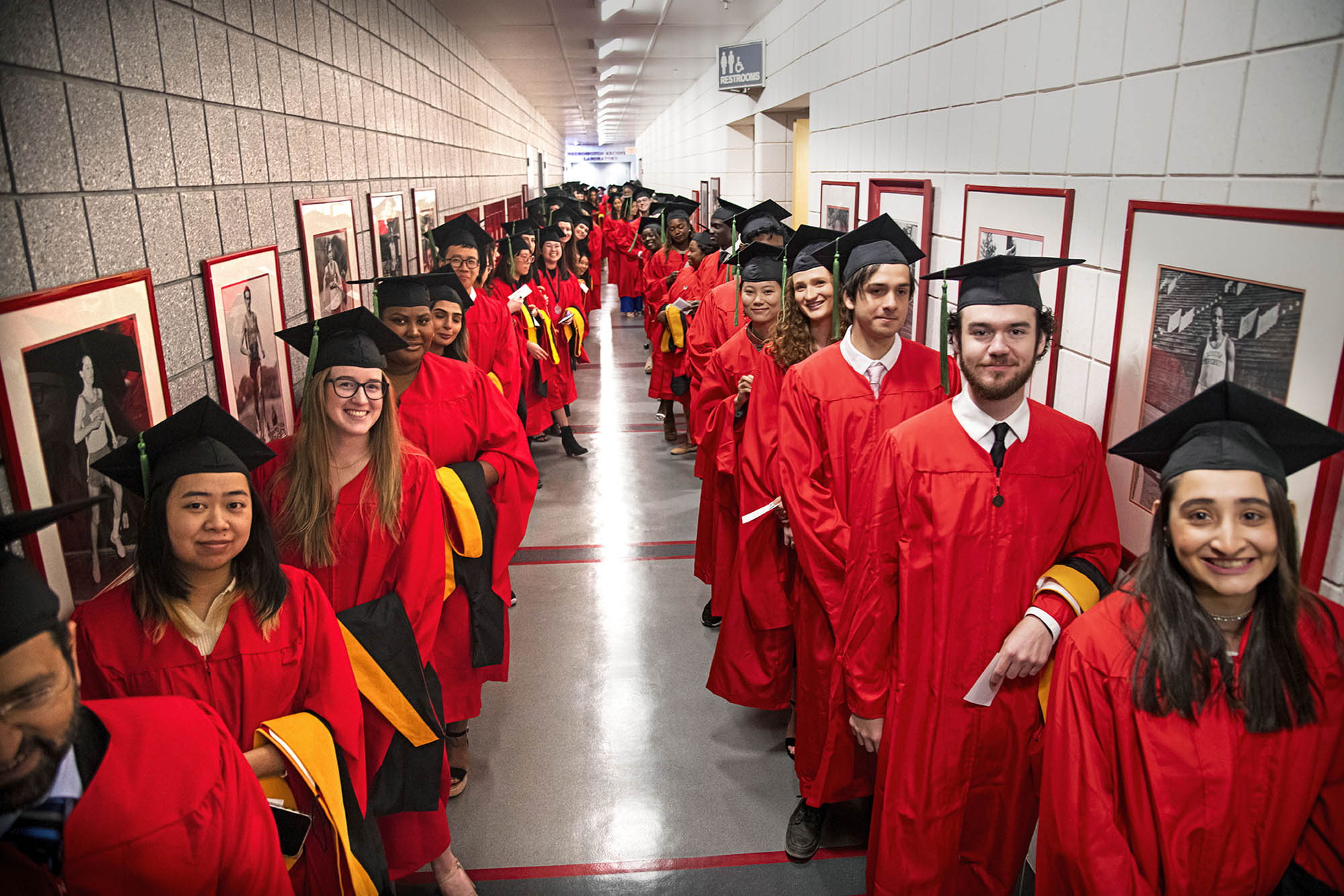 Students wearing red graduation gowns and black graduation caps stand in two parallel lines in a long hallway as they smile and look at the camera. They all hold black and gold stoles in their arms.