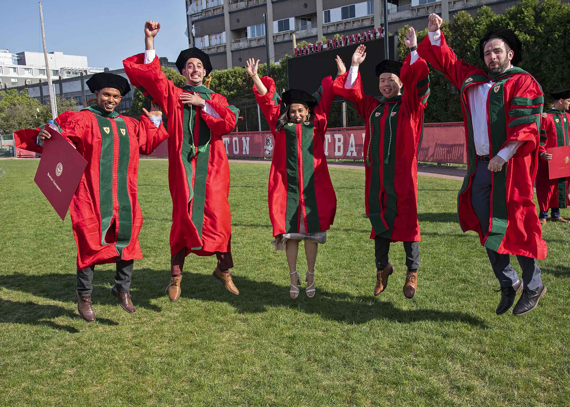 Photo: Five students wearing red graduation gowns, dark green stoles, and black graduation caps laugh as they are cause jumping mid-air.