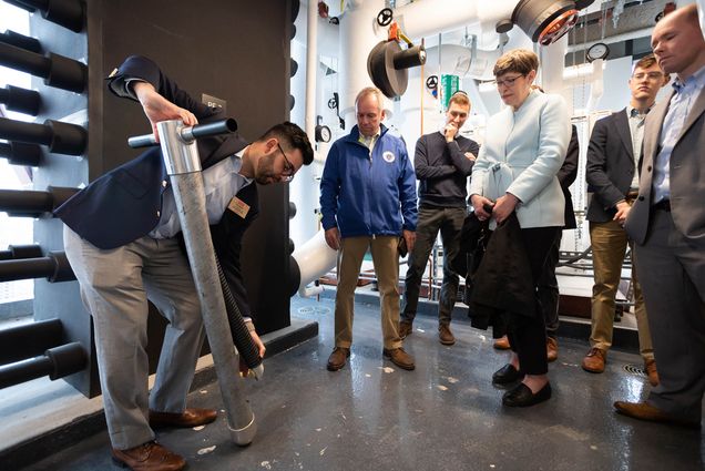 Photo: Sam Moller, BU Assistant Director for Communications (left) demonstrates with the aid of a small scale replicated pipe how the buildings HVAC system obtains itÕs energy. (from left to right) State Representative Jeff Roy (D-Franklin), Emerson Gagnon, State Representative Joan Meschino (D-3rd District Plymouth), Liam Morehead, and State Representative Rich Haggerty (D-30th Middlesex District) look on in the facilities room.