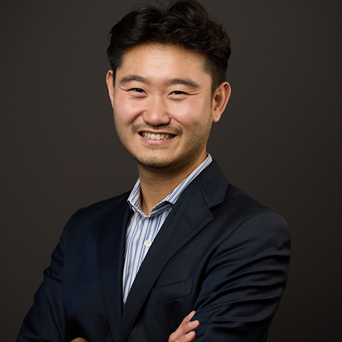 Photo: Charles Chu, an Asian man wearing a light blue collared shirt and navy blue blazer, smiles and poses with arms crossed in front of a dark background.