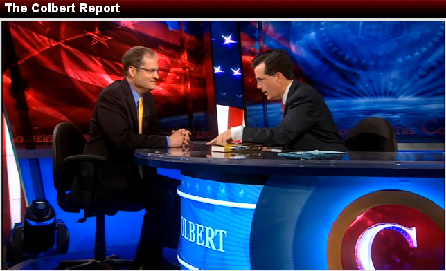 Screenshot of Prothero, a white man wearing glasses, a gold tie, and a black suit seated in front of a large desk as Stephen Colbert, a white man wearing glasses, a red tie, and black suit, and leans toward him and talks. Behind them, a large screen shows a large American flag.
