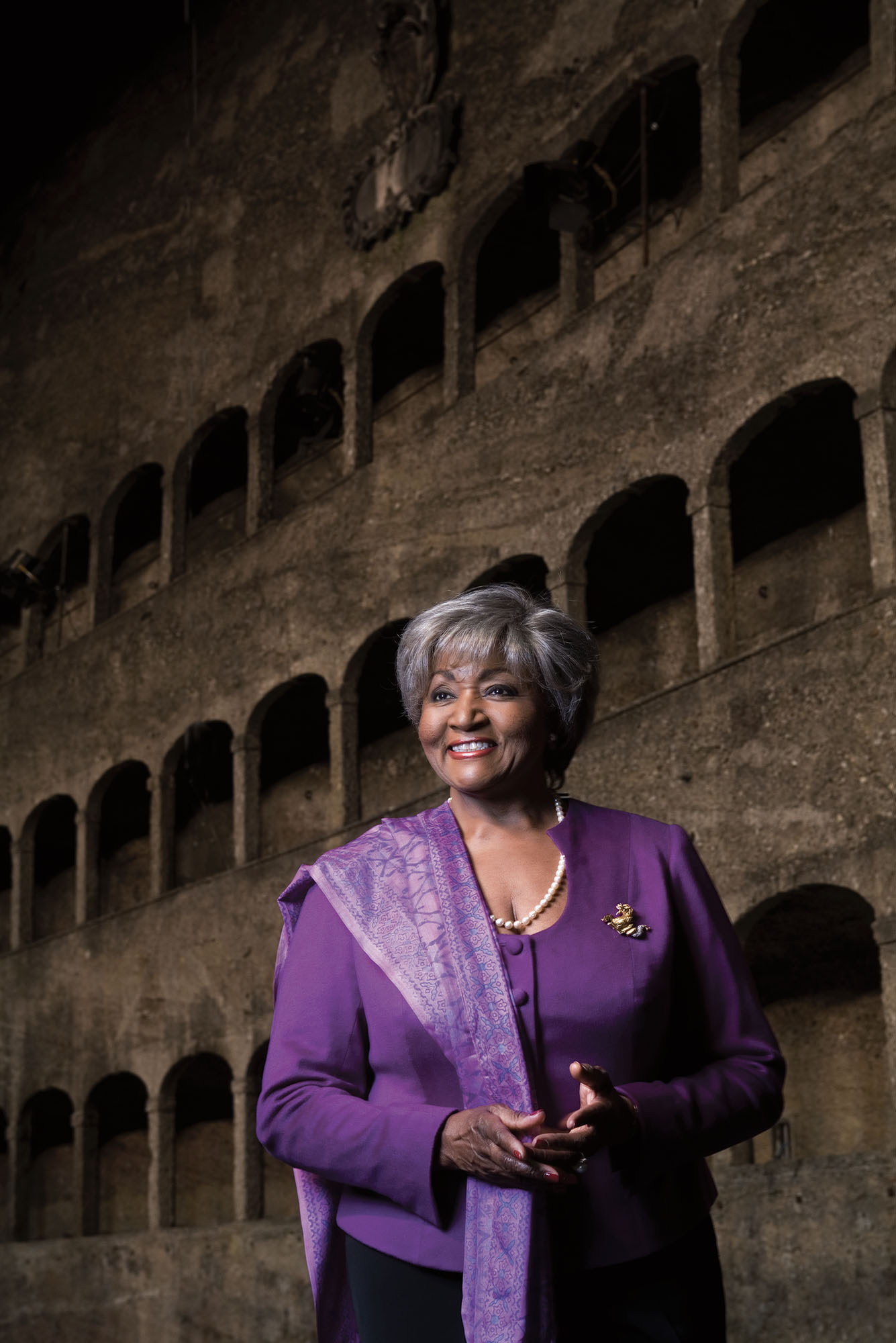 Photo: Grace Bumbry, a Black woman with short gray hair, wears a purple balzer and blouse with a scarf draped over her left side. On her right lapel, a silver brooch. She smiles to the left, big and bright,