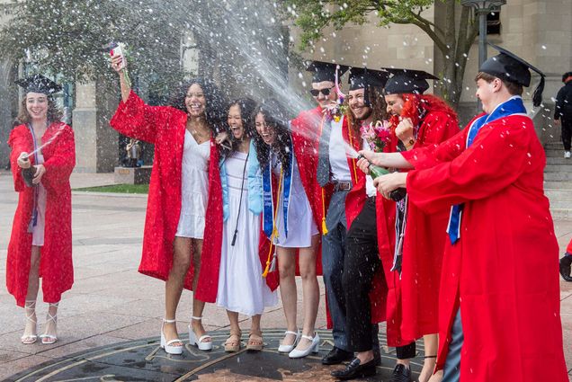 Photo: A group of students wearing BU scarlet red graduation gowns, black graduation caps, and formal wear laugh and smile as they stand on the BU Seal and spray newly opened bottles of champagnes into the air.