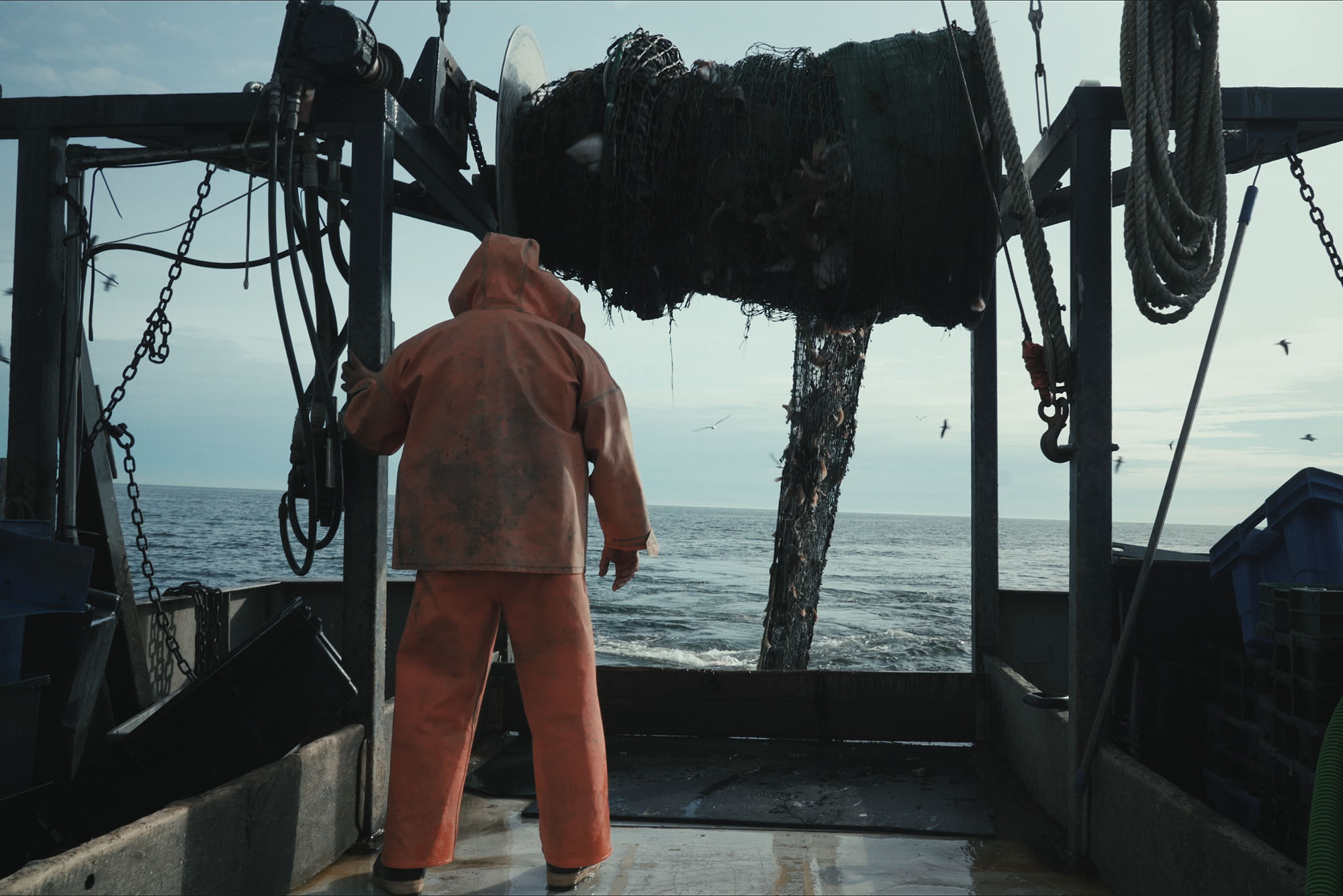 Photo: A cod fisherman in an orange jumpsuit stands on the deck of a fishing boat surrounded by machinery