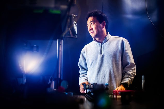 Photo: Engineering Professor, Dr. Ji-Xin Cheng, an Asian man wearing a grey pullover sweater, poses for a photo in his lab. A blue light gives the photo a cool-themed feel as Cheng looks to the left and stands in front of a table full of various mechanical engineering equipment.