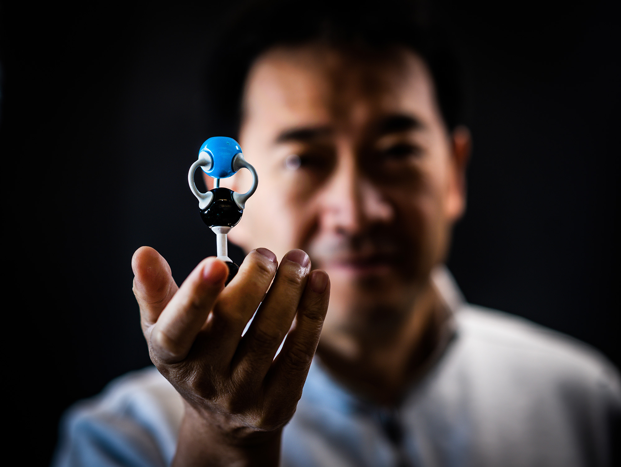 Photo: Engineering Professor, Dr. Ji-Xin Cheng, an Asian man wearing a grey pullover sweater, holds up a small device in the palm of his hand for the camera. The device, a blue ball held up by tiny machinery, is focused in the foreground as Cheng's face and body are blurred in the background.