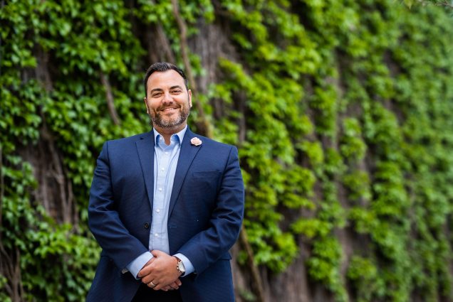 Photo: Jason Campbell-Foster, a white man with dark hair and a slightly graying beard and mustache wearing a light blue collared shirt and navy suit, poses in front of an ivy-covered wall. He stands with hands clasped in front of him as he smiles for the camera.