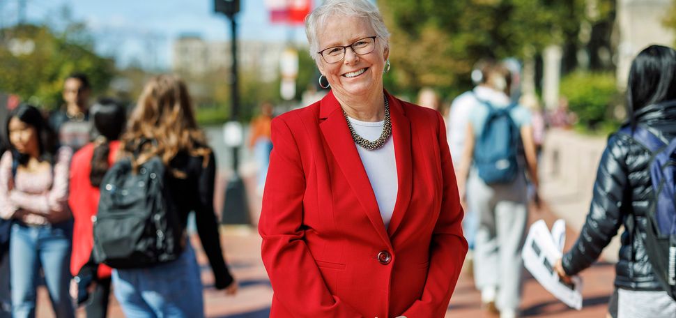 Photo: Portrait of Boston University Provost Jean Morrison on Commonwealth Ave. Asmiling white woman with short, silver white hair and wearing glasses, a white blouse, and red blazer poses with fingers clasped in front of her on a bustling sidewalk. Background and passerby are blurry.