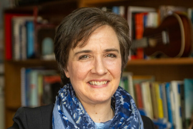 Photo: A white woman with short, brown hair looks into the camera and smiles. She wears a black cardigan and a blue-patterned infinity scarf. Behind her is a stack of books and a guitar.