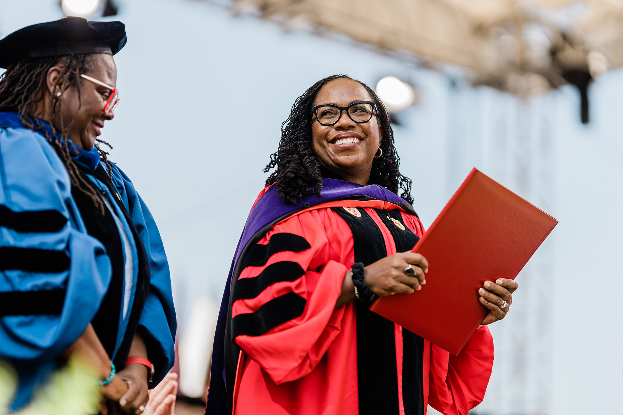 Photo: Ketanji Brown Jackson, a black woman with long sister locs and wearing glasses, a red ceremonial graduation gown, and purple stole, smiles as she holds her honorary degree in her hand. To her left stands a smiling Angela Onwuachi-Willig, a Black woman wearing a blue ceremonial graduation gown and black cap.