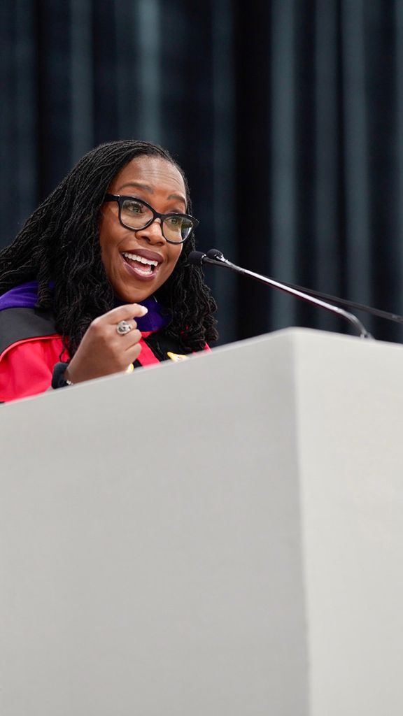 Photo: Ketanji Brown Jackson, a Black woman with long dreadlocks wearing a red ceremonial and purple stole smiles as she speaks into a white Boston University podium.