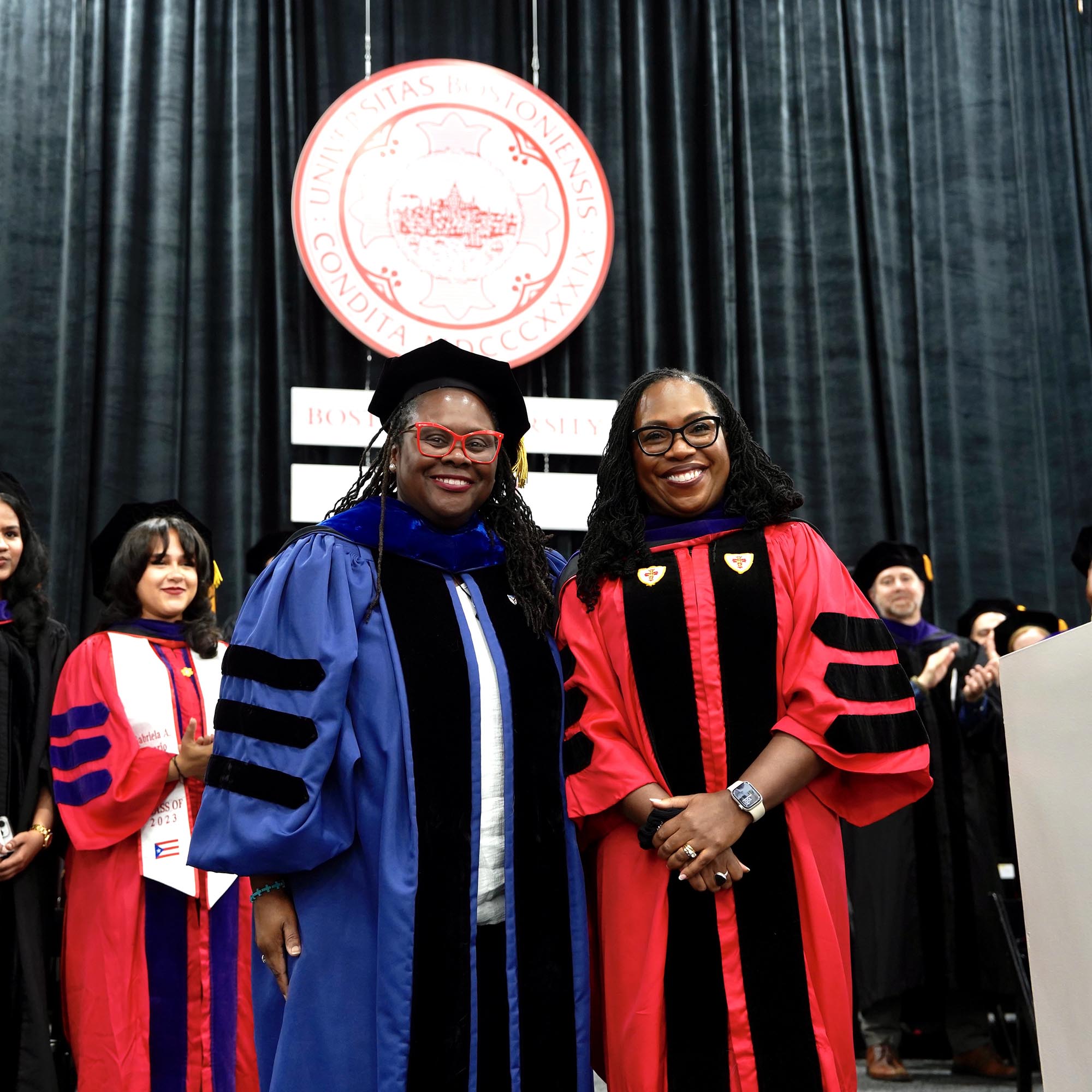 Photo: Angela Onwuachi-Willig, a Black woman wearing a blue ceremonial graduation gown, black cap, and black stole (left), smiles and poses for a photo with Ketanji Brown Jackson, a Black woman with long dreadlocks wearing a red ceremonial and purple stole.