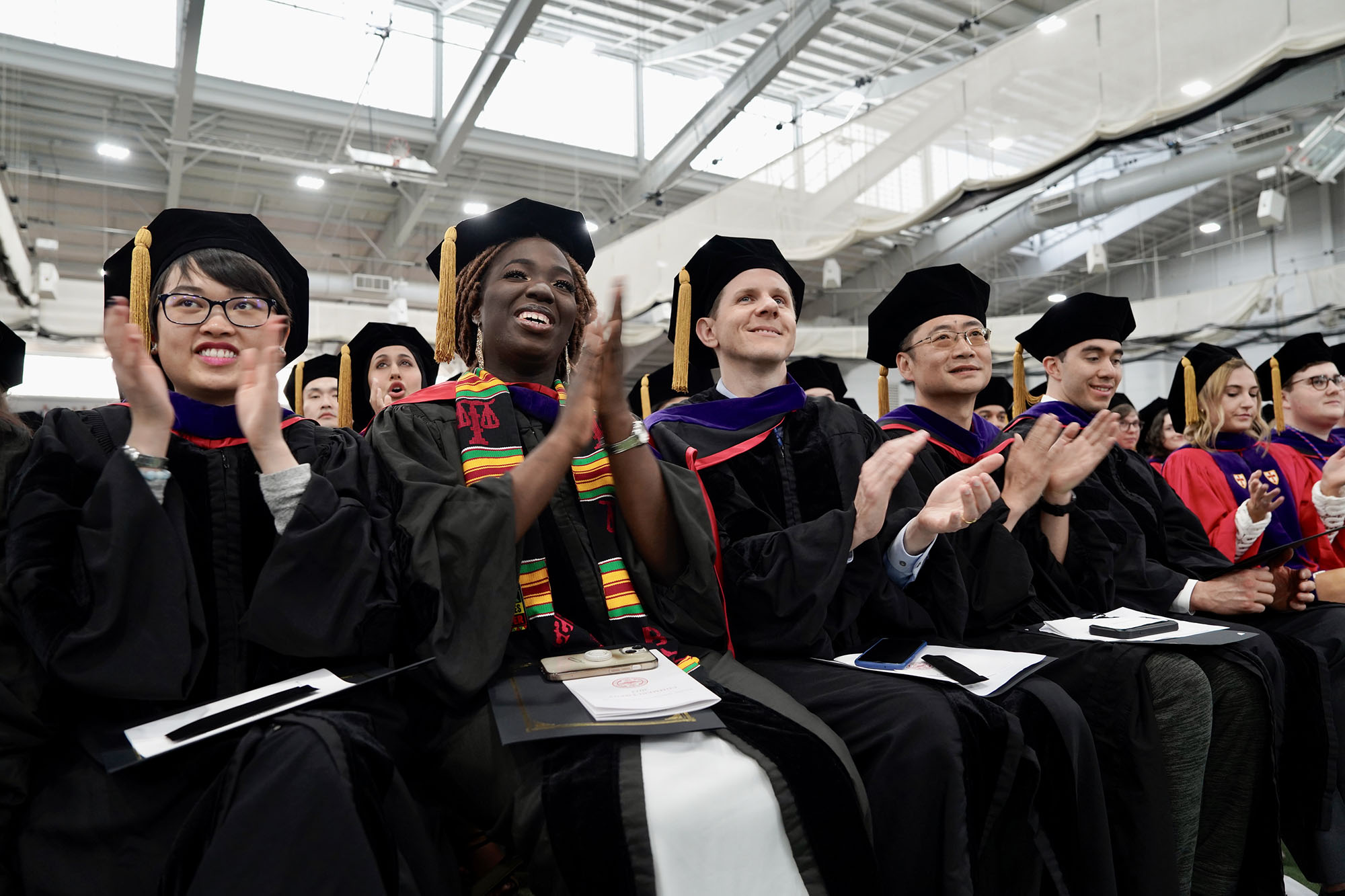 Photo: Students sitting and wearing black graduation gowns and caps clap and smile as they look towards the stage.