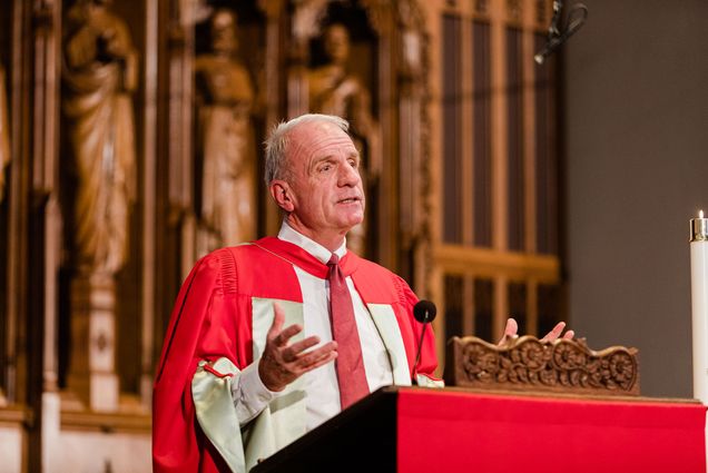 Photo: Rev. Robert Allan Hill, a white man wearing white and red ceremonial wear, stands with hands raised palm-up in way of prayer, as he speaks at a wooden podium in Marsh Chapel.