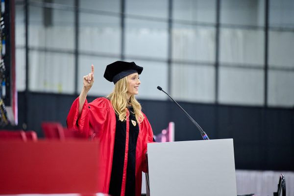 Photo: Kim Raver stands at a podium wearing a red robe and black masters cap while addressing a large crowd at the College of Fine Arts convocation at Boston University in May 2023