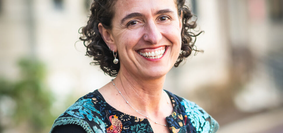 Photo: CAS professor Joanna Davidson, a white woman with short, curly brown and wearing a blue mosaic-themed blouse and black long-sleeved top smiles and poses with arms clasped behind her back. A blurry outdoor BU building can be seen behind her.