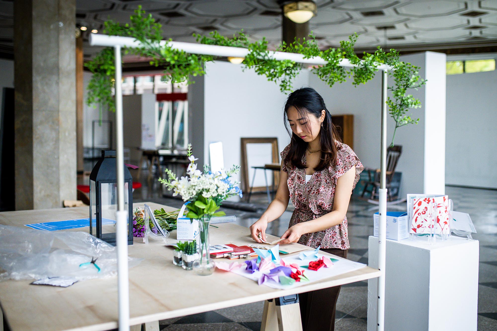 Photo: Mina Chung, a young Asian woman wearing a pink floral blouse and brown pants, stands and works under a green canopy on a table.