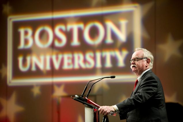Photo: President Brown, a white man with white hair and a thick, white mustchae, stands at a podium with two mics directed at him. To his right and behind him, there is a light production with BOSTON UNIVERSITY with stars in the background.
