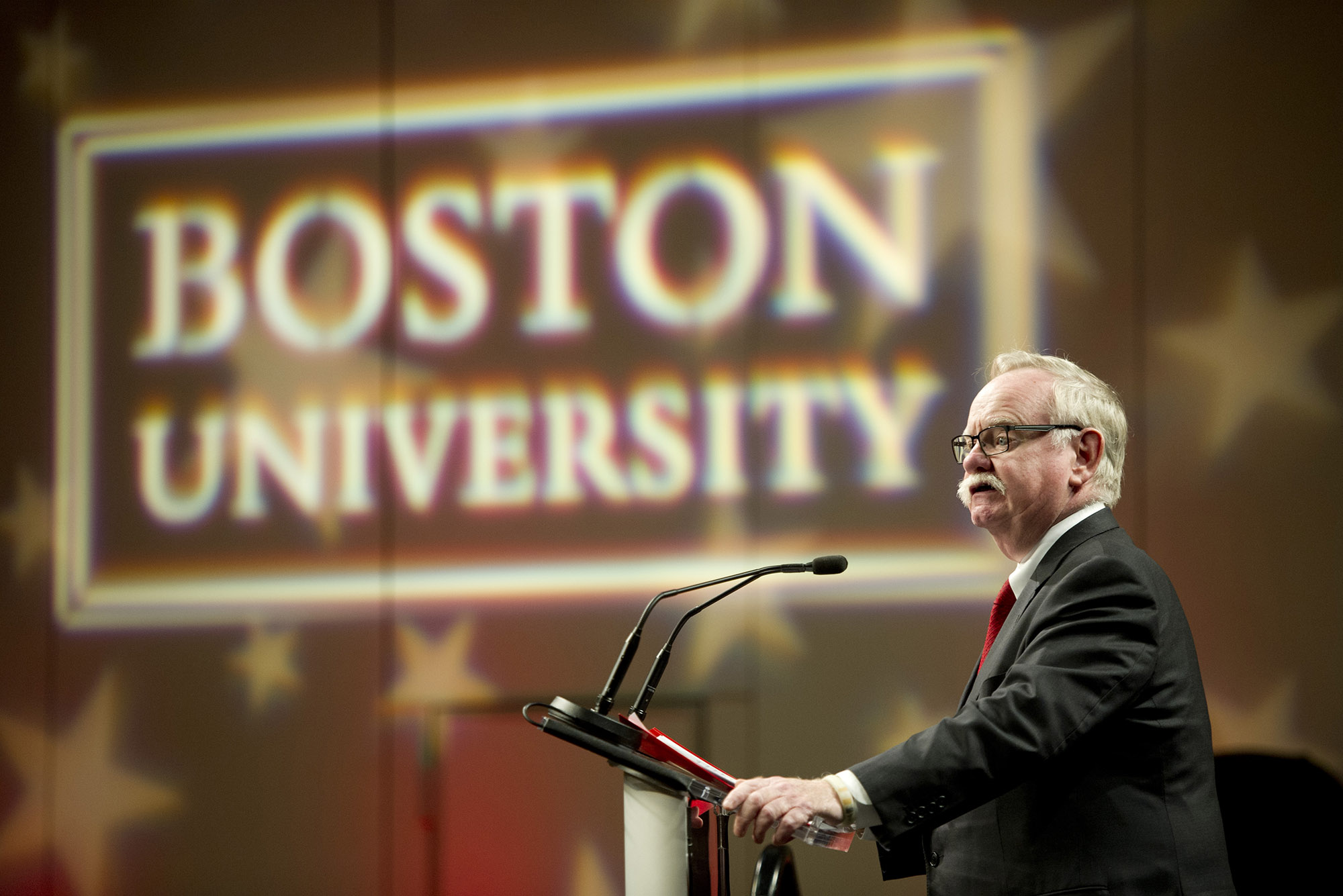 Photo: President Brown, a white man with white hair and a thick, white mustchae, stands at a podium with two mics directed at him. To his right and behind him, there is a light production with BOSTON UNIVERSITY with stars in the background.