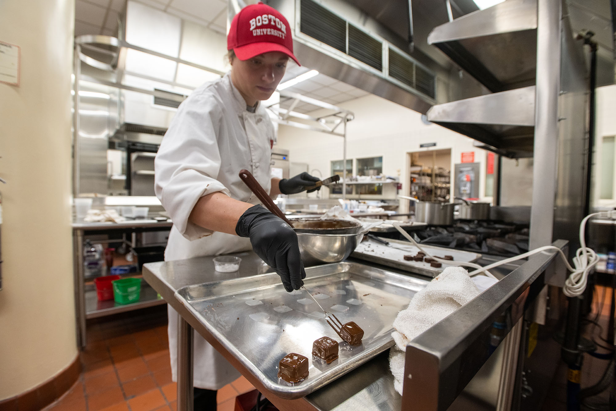 Photo: Mia Ball, a person wearing a red BU cap and white chef's outfit, dips cubes of caramel in tempered chocolate as they stand in a large test kitchen.