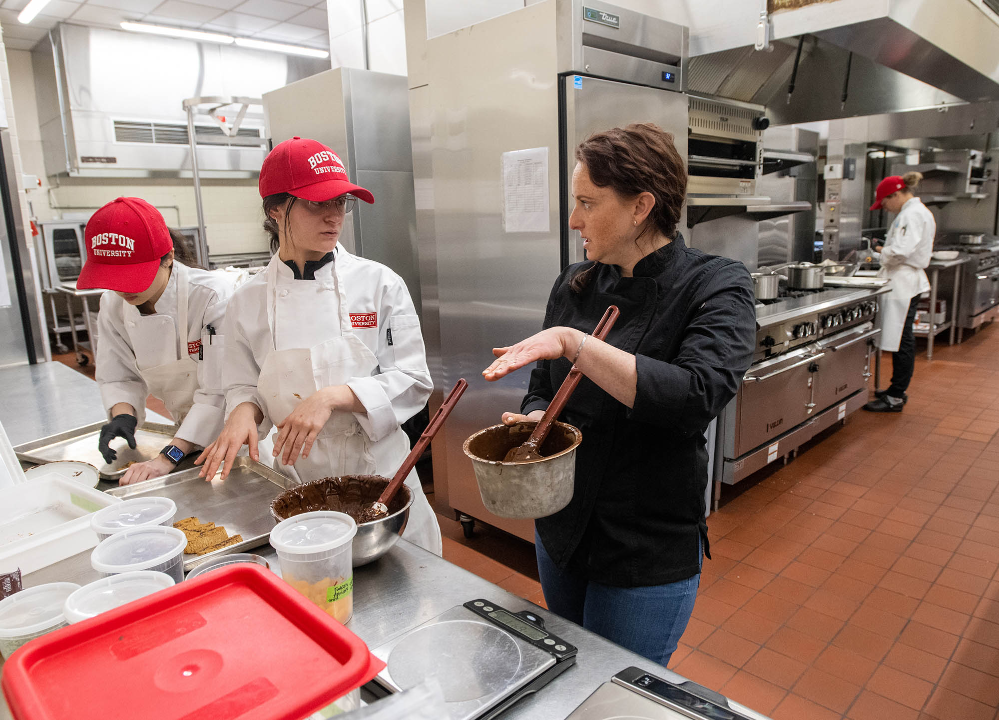 Photo: Mara Clement, a young person wearing a red BU cap and white chef's outfit, with chef instructor Anne Wolf, a woman wearing a black long-sleeved chef top and jeans, during the chocolate work class during the Professional Program in Pastry Arts March 30. Wolf holds up a large pot of chocolate as she explains something to Clement.