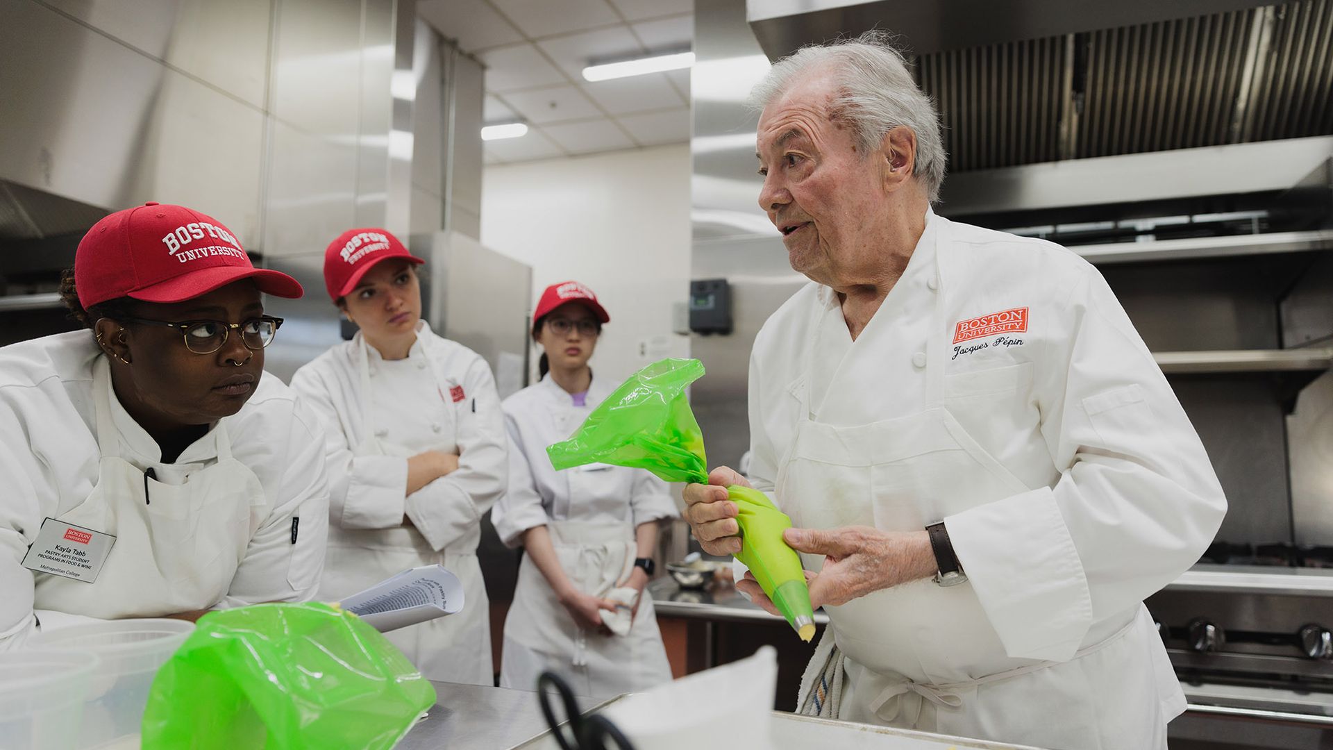 Photo: 3 students look on as renowned chef Jacques Pépin, an older white man with silver white hair and wearing a white chef's jacket with his name sewn onto the breast pocket, holds a green piping bag and speaks to the students.