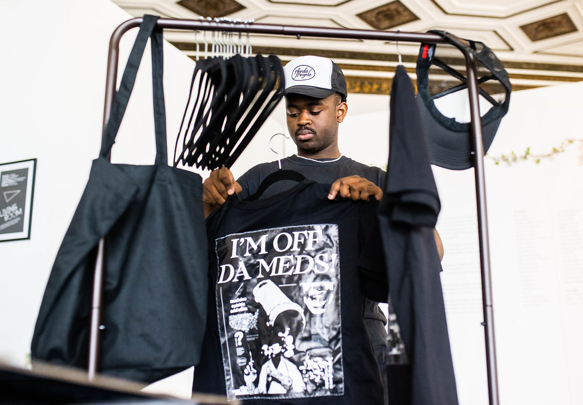 Photo: A young Black man wearing a black and white baseball cap sort and arranges printed totes and t-shirts on a metal garment rack.