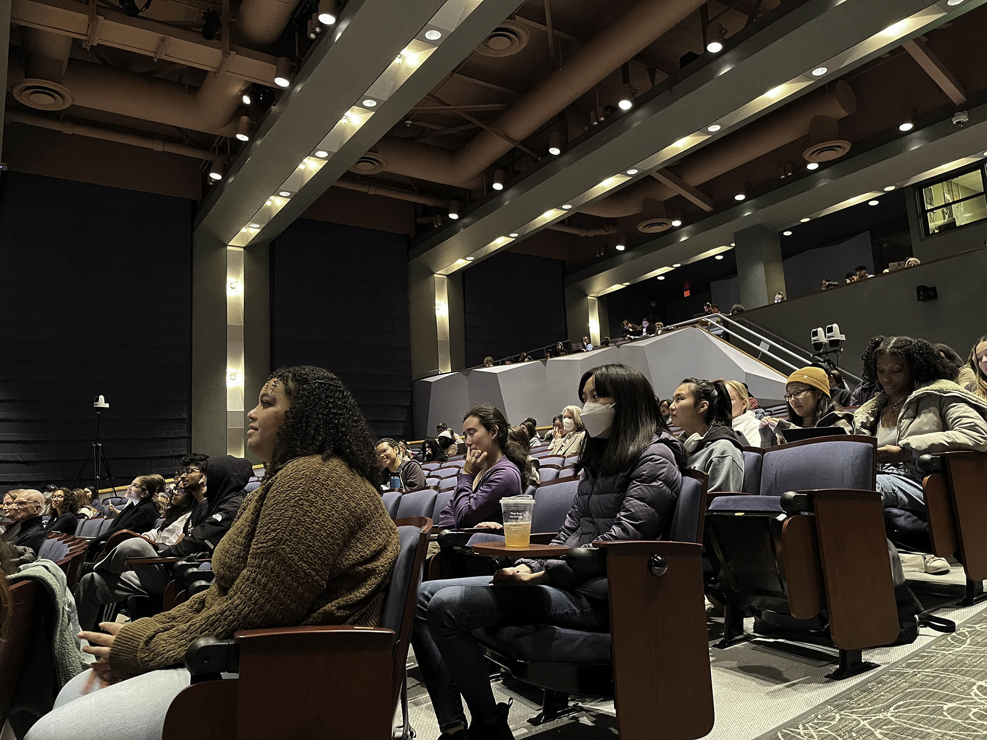 Photo: A large lecture hall is shown filled with students looking to the front of the room attentively.