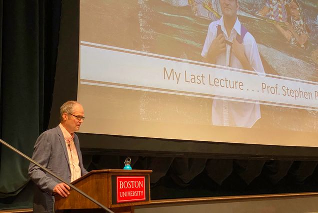 An older white man with dark gray hair and wearing glasses, a cream-colored collared shirt and grey blazer, reads from a podium as a large screen behind him displays a powerpoint that reads "My Last Lecture".