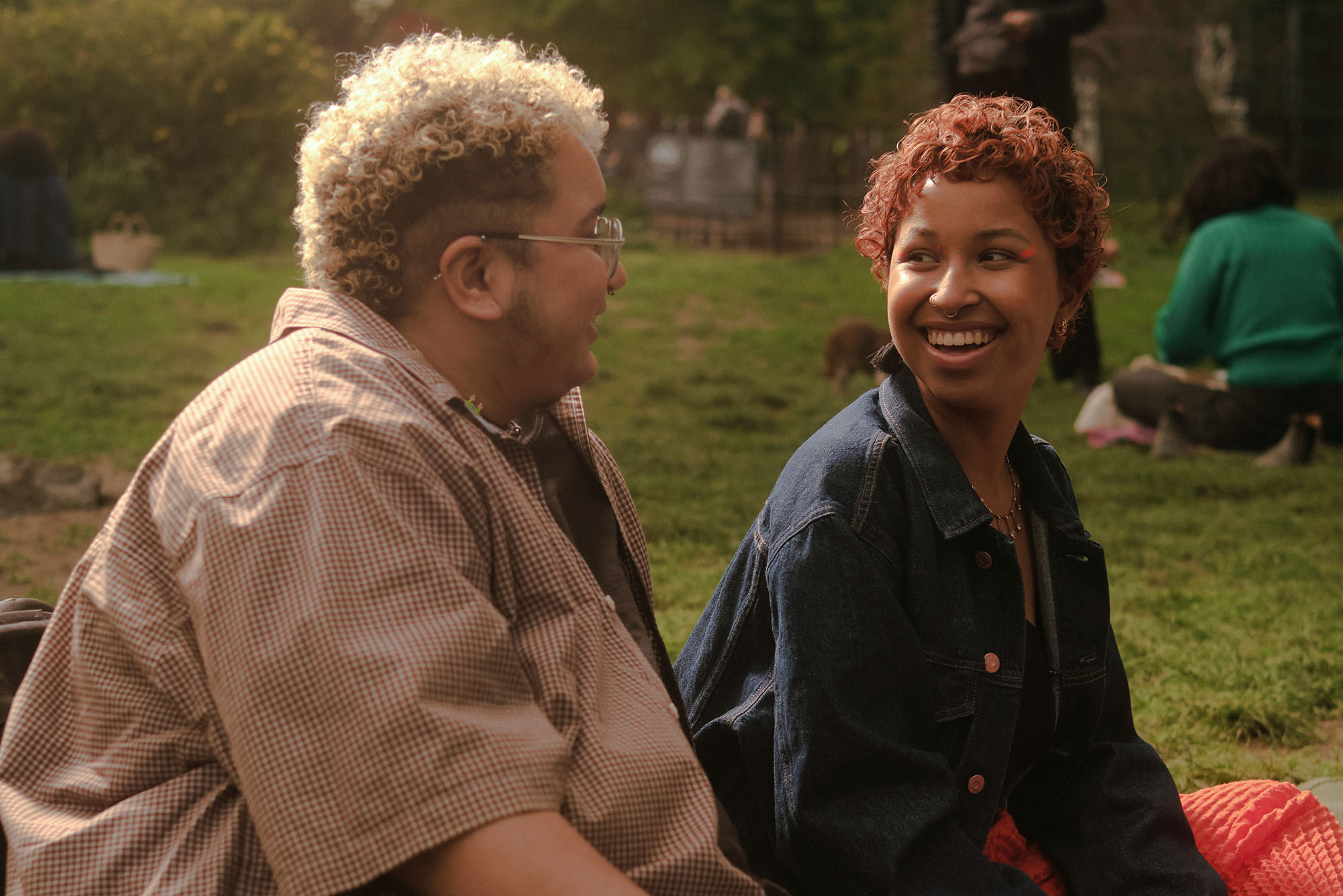 Photo: A trans couple laugh and looks at each other lovingly in the golden hour of sunlight. They lounge on a blanket in a park.