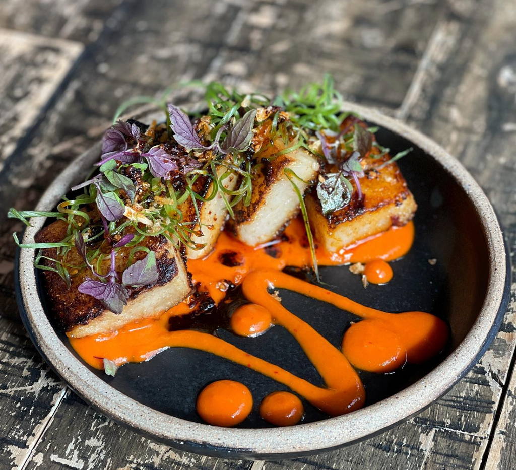 This is a brunch dish served at the restaurant Little Donkey in Cambridge. The food is on a black plate and there is sliced chicken with an orange sauce and micro greens for garnish.